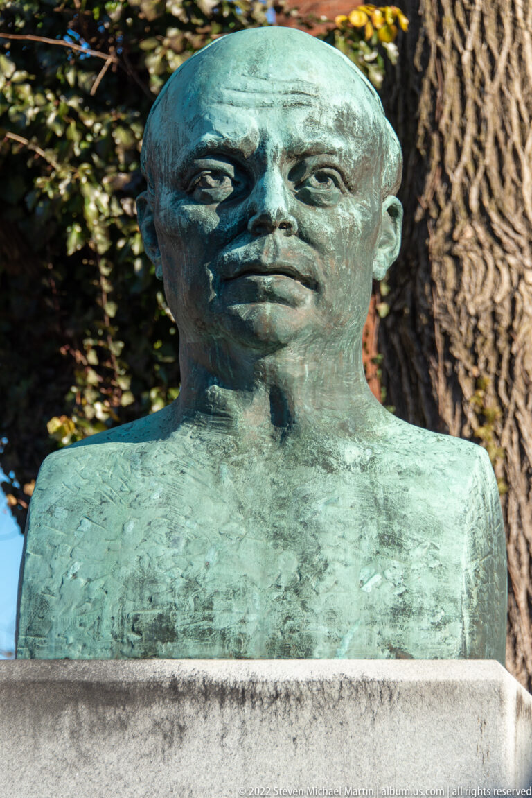 A weathered bronze bust of a suited man stands on a stone pedestal outdoors.