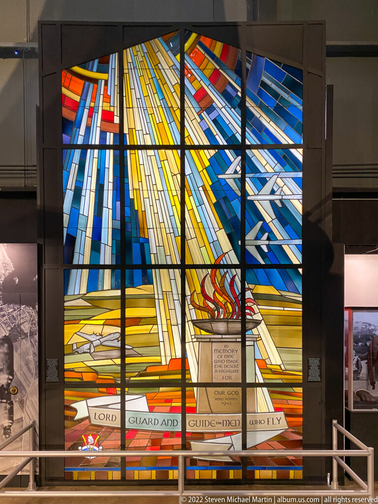 A blue, yellow, and red stained-glass panel includes a flame motif and a prayer ribbon.