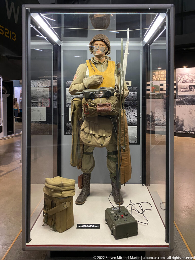 A uniformed figure with a large pack and radio equipment stands in a glass case beside archival panels.