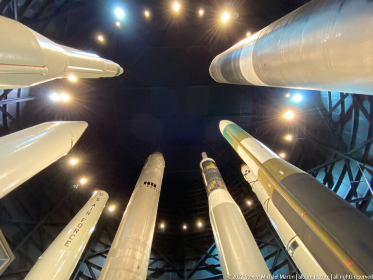 Long missiles and rockets hang overhead, their pointed noses converging above the viewer.