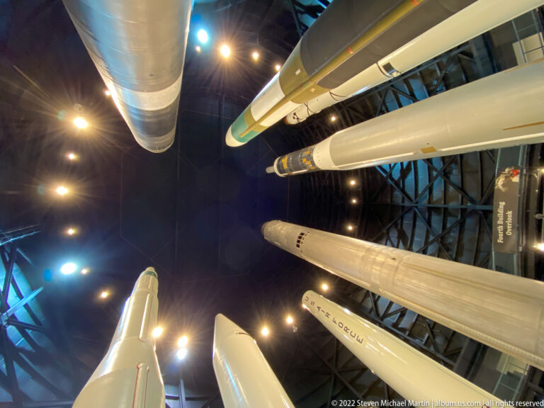 Multiple long rockets and missiles cross overhead beneath spotlights in a dark gallery.