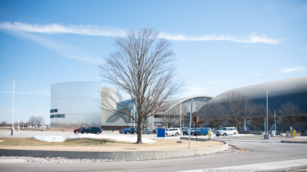 Curved-roof museum hangars and a cylindrical building stand beside a winter road and parking area.