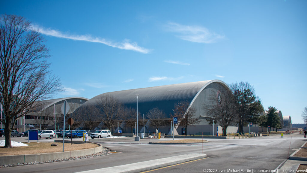 Two large curved-roof museum hangars line a campus road beside a bare tree and parked cars.
