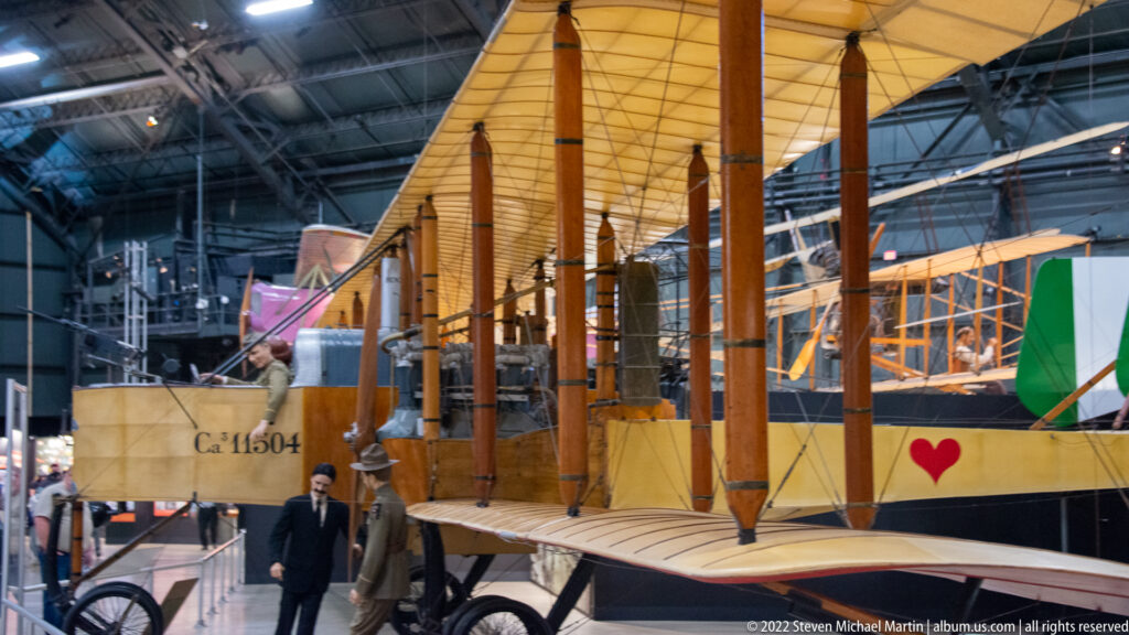 A yellow biplane with wooden struts and a red heart marking stands beside a visitor.