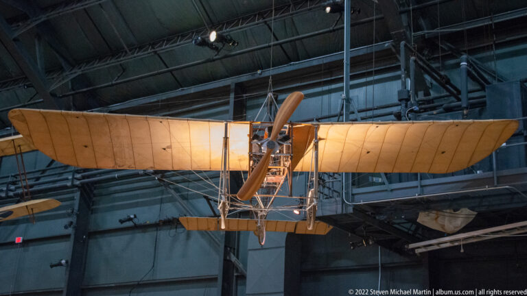 A tan early biplane hangs overhead, exposing its stacked wings, propeller, struts, and wires.