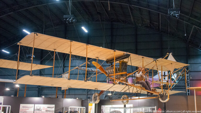 A suspended tan biplane has an open cockpit with a seated figure beneath its long wings.