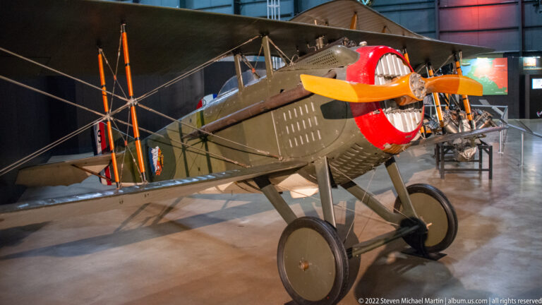 An olive biplane with an open cockpit and red-and-white circular nose marking stands on spoked wheels.