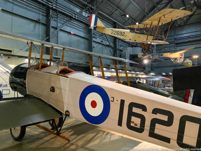 A cream biplane with a blue-white-red roundel and number 1626 stands beneath another aircraft.