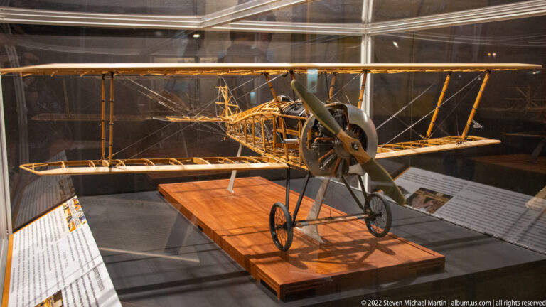 A small early biplane with wire-braced wings and narrow landing gear stands inside a glass case.