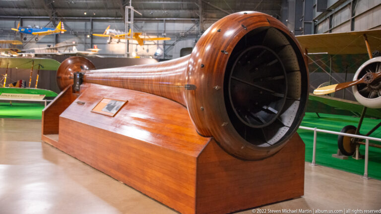 A polished wooden cylinder with a wide dark opening sits on a display base among aircraft.