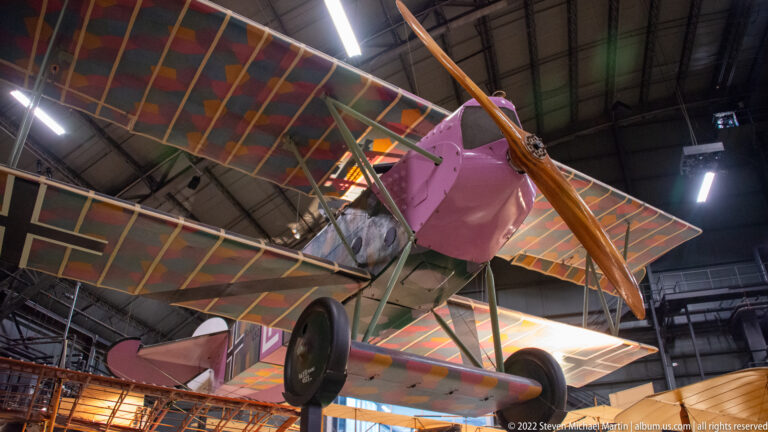 A colorful pink biplane hangs overhead, showing its wheels, struts, propeller, and patterned wings.