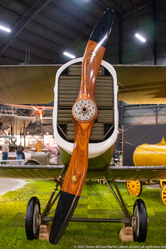 A dark wooden propeller fills the foreground of a small aircraft, with its nose and landing wheels behind.