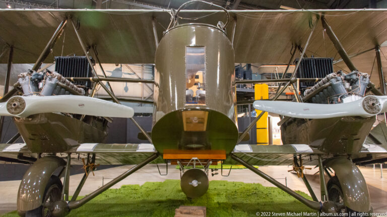 An olive-drab aircraft faces forward with a central cockpit, paired guns, and broad landing gear.