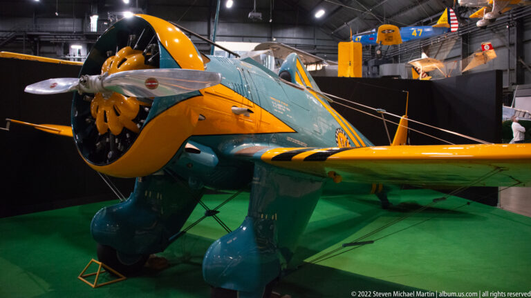 A blue-green aircraft with yellow wings and rounded wheel fairings stands on a green museum floor.