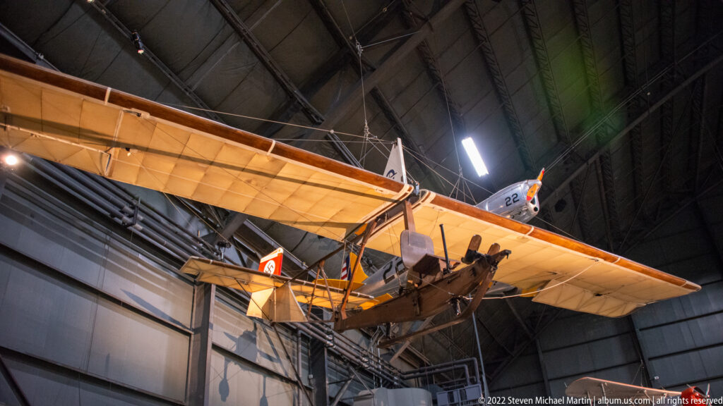 A tan biplane hangs beneath the museum roof, with its wings, open cockpit, and landing gear visible.