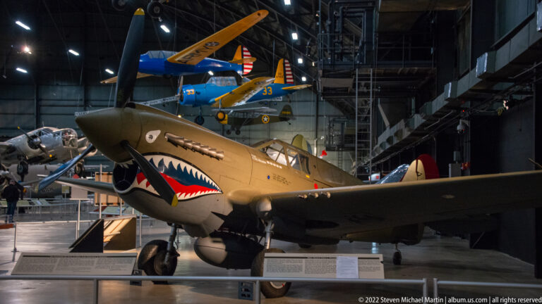 A tan fighter aircraft with shark-mouth nose art stands beneath a blue plane in the museum hangar.