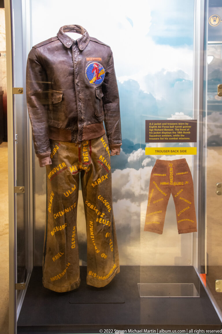 A leather flight jacket with a shoulder patch and decorated trousers stand in a glass case.