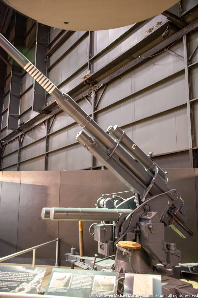 A large metal aircraft gun assembly with a long barrel and angular supports stands above interpretive panels.