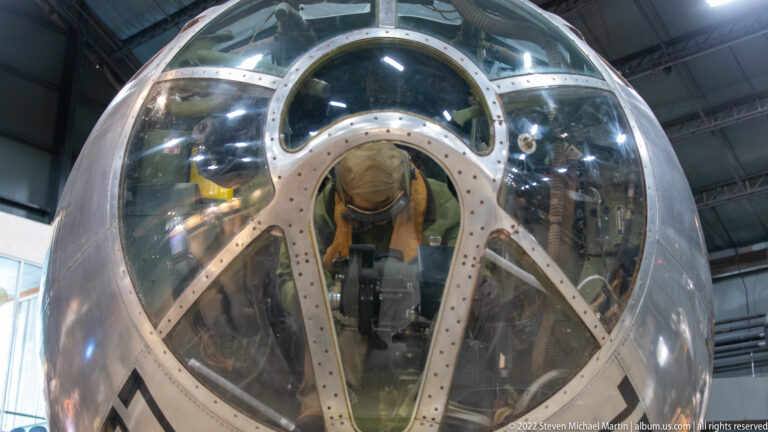 A mannequin sits inside a B-17’s rounded glazed nose, surrounded by curved metal framing.
