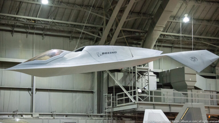 A sleek white aircraft marked BOEING sits on a raised stand beneath the museum’s steel trusses.