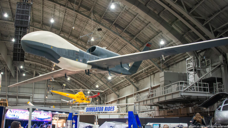 A large white and blue transport aircraft is displayed overhead in a hangar, above a yellow aircraft and museum exhibits.