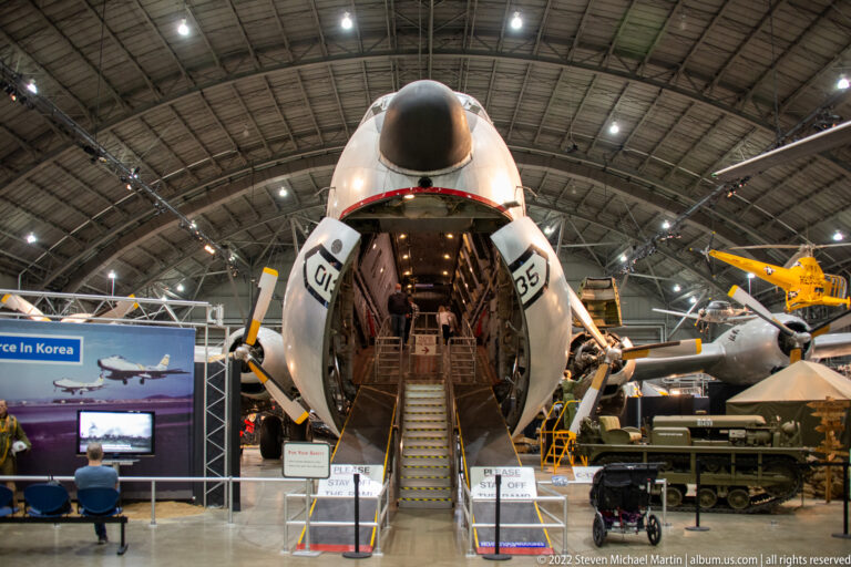 A transport aircraft stands with its nose raised, exposing the cargo bay and a stairway, with exhibit barriers in front.