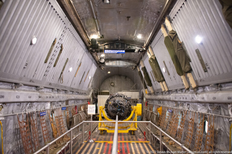 A long aircraft cargo compartment has ribbed metal walls, visitor rails, and a mechanical assembly displayed at its center.