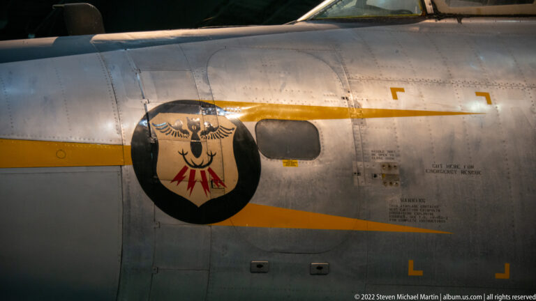 A black shield emblem and yellow stripe cross a silver aircraft fuselage with windows, rivets, and small stenciled markings.