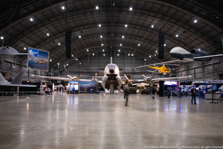 Visitors walk through a broad aircraft hangar with planes and exhibit panels arranged beneath a high, brightly lit roof.