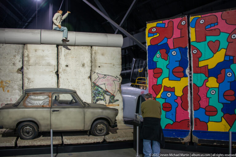 A compact gray car stands beside a graffiti-marked wall and colorful panels, while a visitor examines the display.