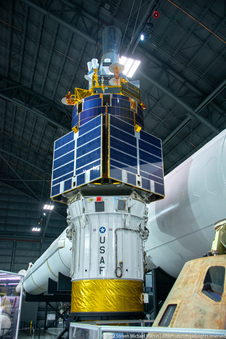 A U.S. Air Force satellite with blue solar panels and gold details is displayed above a platform inside the museum hangar.