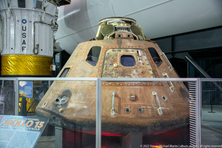 The Apollo 15 command module sits behind a clear barrier, showing a scorched conical shell and fittings beside a white-and-gold module.
