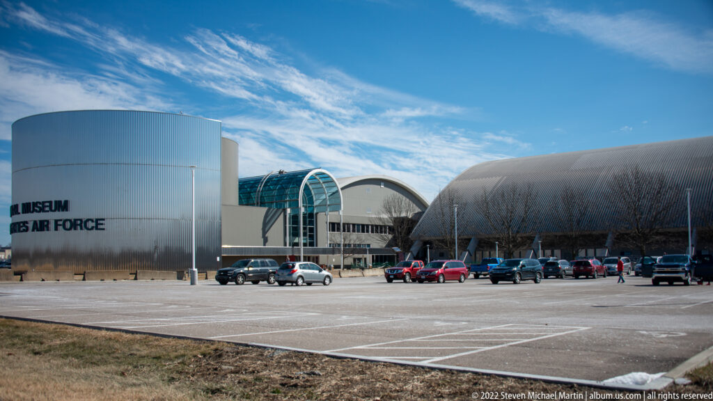 A cylindrical museum entrance tower with the institution’s name stands before curved metallic halls and a parking lot under a blue sky.