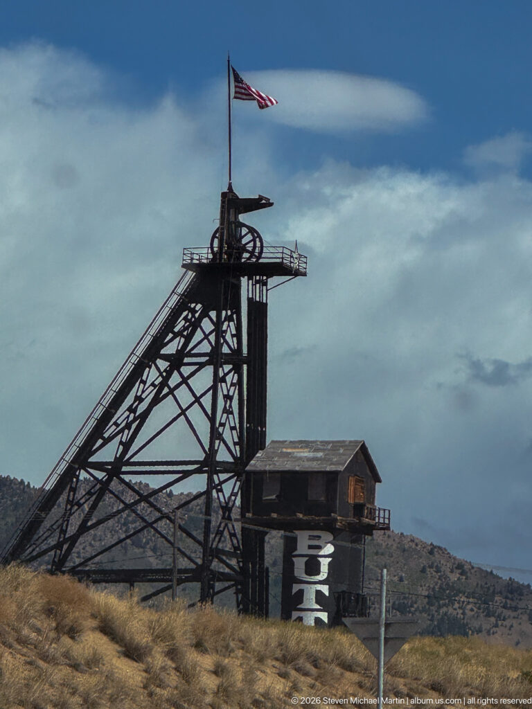 Close side view of black steel mine headframe with American flag and adjacent dark building marked BH.