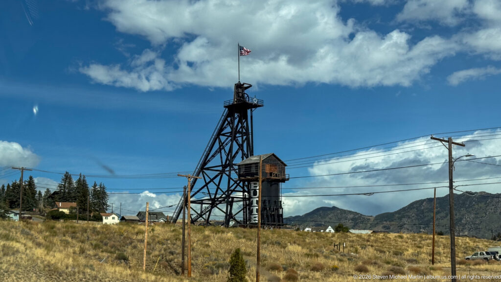 Black steel mine headframe with American flag, adjacent shaft structure, utility lines, dry grass, and mountains.