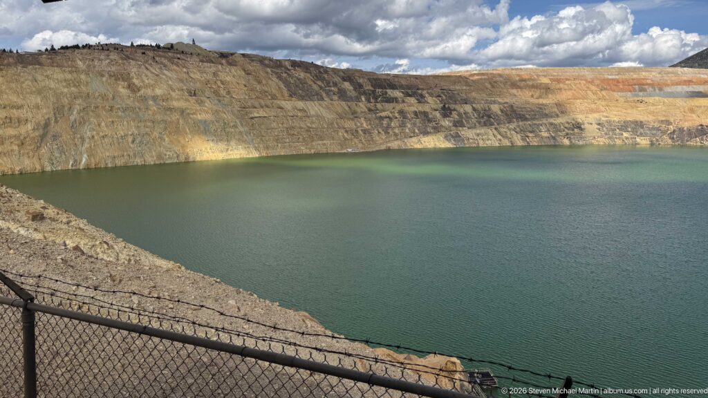 Wide green Berkeley Pit lake below layered gray, ocher, and rust-colored mine benches under broken clouds.