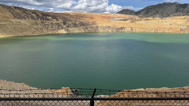 Dark teal Berkeley Pit water beneath sunlit yellow and orange mine benches, with a fence along the foreground.