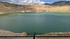 Dark teal Berkeley Pit water beneath sunlit yellow and orange mine benches, with a fence along the foreground.