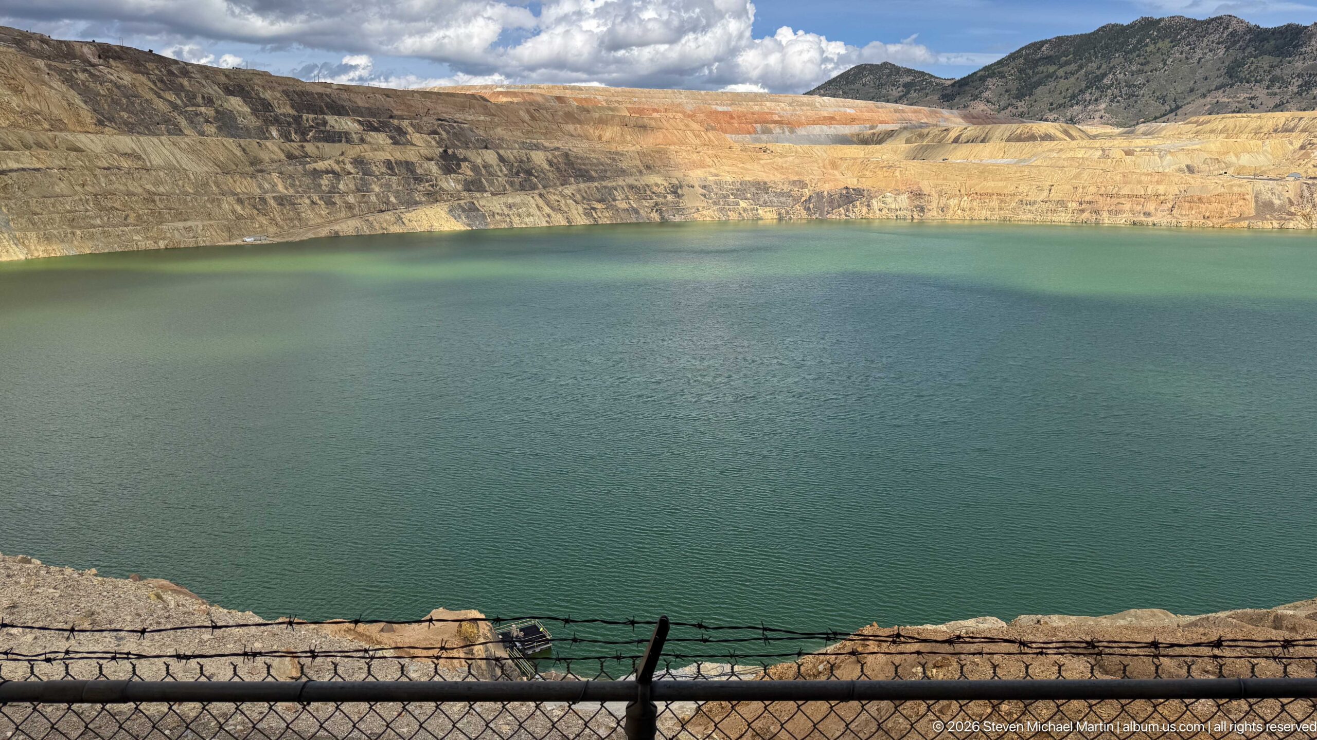 Dark teal Berkeley Pit water beneath sunlit yellow and orange mine benches, with a fence along the foreground.