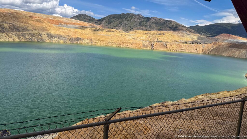 Close view across green-blue Berkeley Pit water toward yellow and rust-colored terraced mine walls and dark mountains.