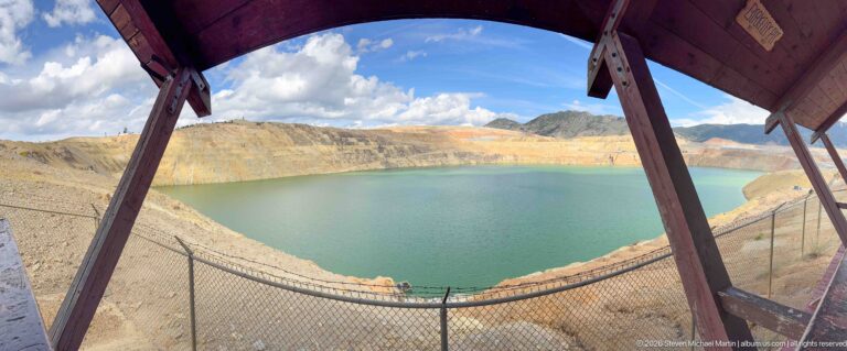 Green-blue Berkeley Pit lake surrounded by terraced mine walls, framed by timber supports and a safety fence.