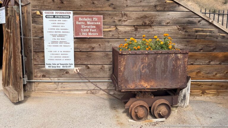 Rusty steel mine cart on four flanged wheels beside a timber wall with Berkeley Pit and Butte signs.