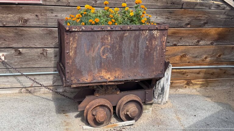 Rusty rectangular steel mine cart on four small wheels beside a weathered timber wall and orange flowers.