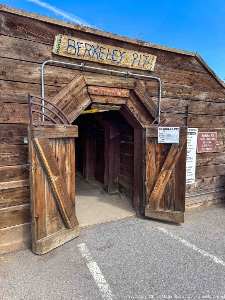 Open double timber doors beneath a Berkeley Pit sign leading into a dark mine-themed entrance passage.
