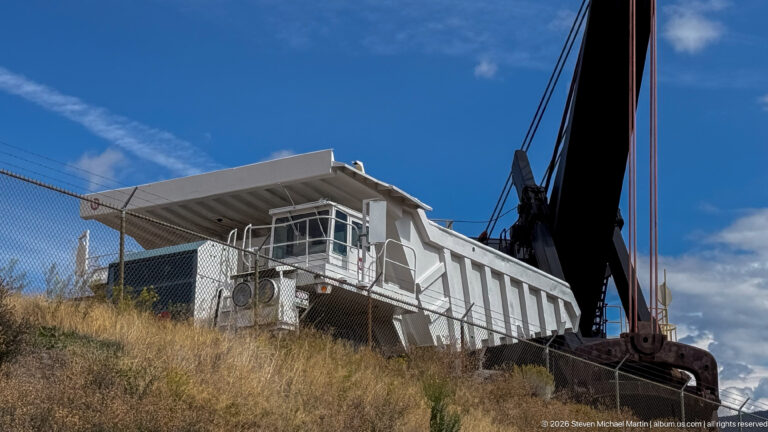 Large black mining machine with heavy cables and pulleys beside an elevated white operator’s cab on a grassy slope.