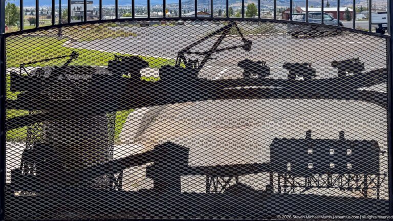 Large black mining machinery with drums, wheels, and shafts displayed outdoors behind protective metal mesh.