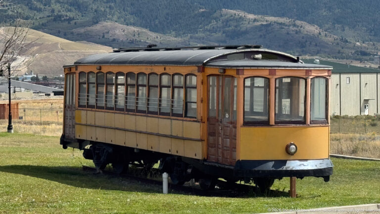 Preserved yellow-brown wooden streetcar with rows of windows standing on short track in an open grassy area.