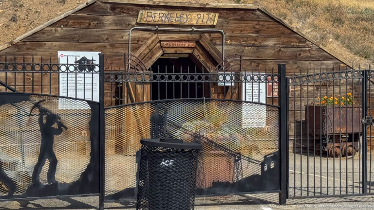 Timber entrance marked Berkeley Pit behind black metal fencing with decorative mining silhouettes and interpretive panels.