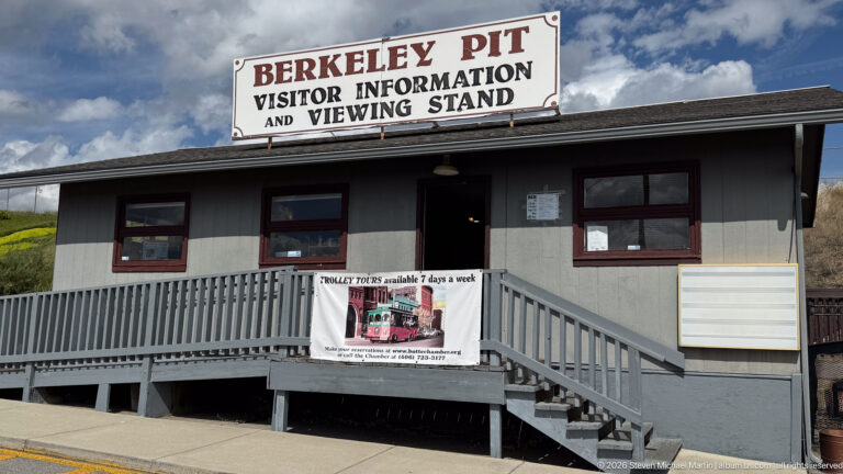 Gray visitor building with rooftop sign reading Berkeley Pit Visitor Information and Viewing Stand, with stairs and ramp.