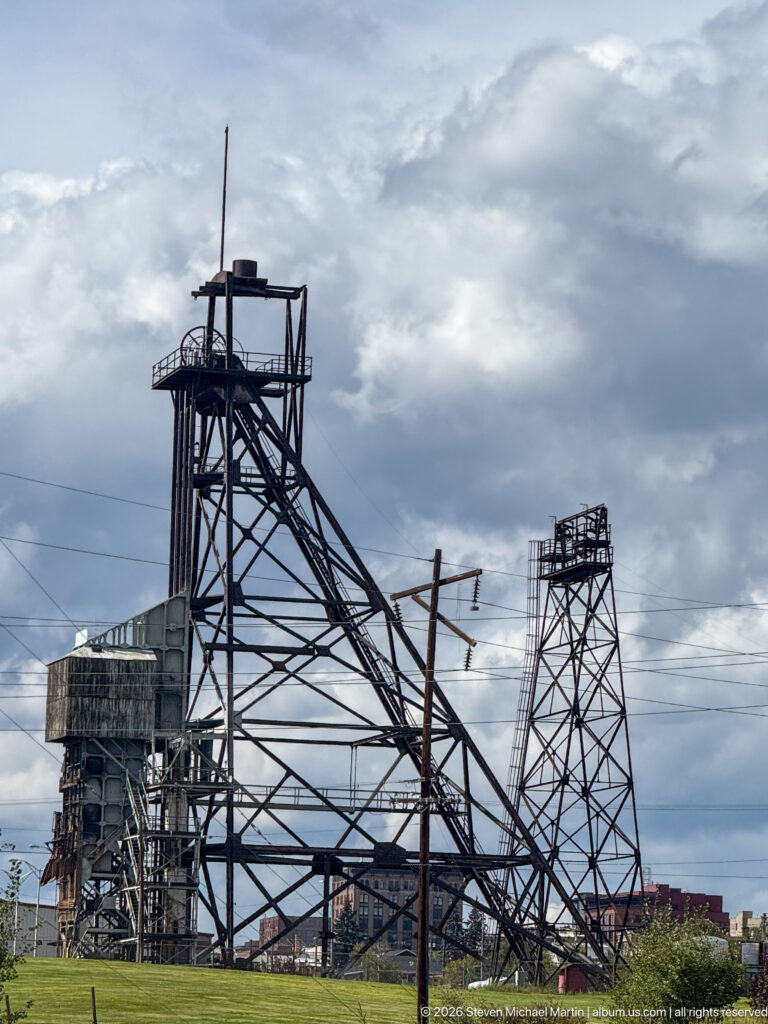 Two tall steel mine headframes and associated industrial structures silhouetted against a cloudy sky in Butte.